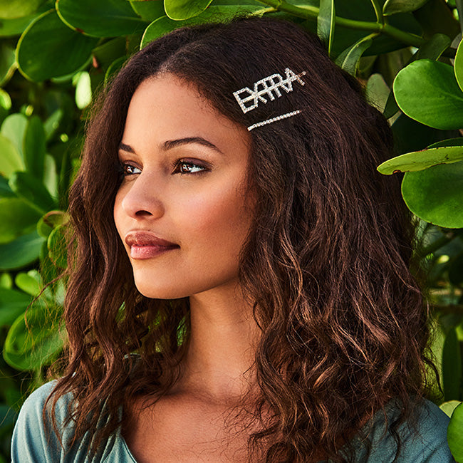 Woman with long, wavy brown hair shown in profile wearing a rhinestone “Extra” barrette, with green leaves behind her