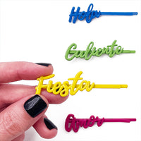 Hand with black nail polish holding a yellow “Fiesta” Atrevida barrette, above blue “Hola,” green “Caliente,” and pink “Amor” barrettes
