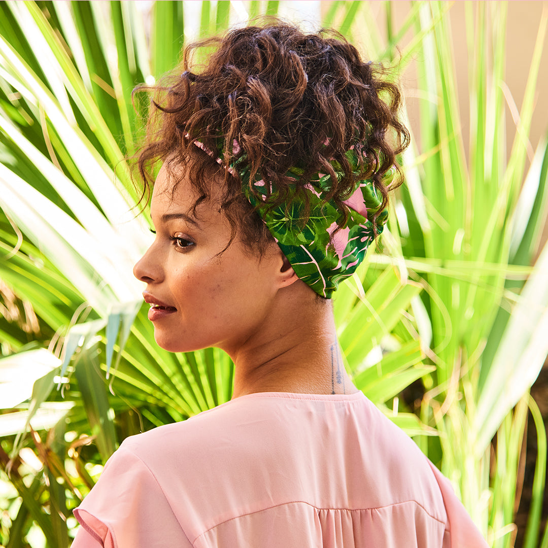 Back view of a woman wearing the tropical leaf print headband around her gathered curls, with palm leaves behind her