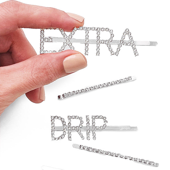 Hand with nude nail polish holding a rhinestone “Extra” barrette, above a rhinestone “Drip” barrette and two straight rhinestone barrettes
