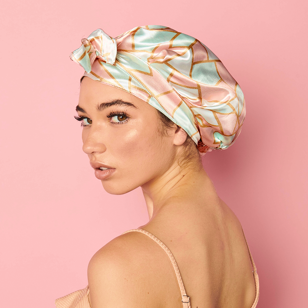 Woman looking over her shoulder against a pink background, wearing the Adjustable Satin Bonnet in a pink, mint, and cream geometric print, tied at the front