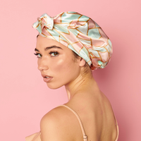 Woman looking over her shoulder against a pink background, wearing the Adjustable Satin Bonnet in a pink, mint, and cream geometric print, tied at the front