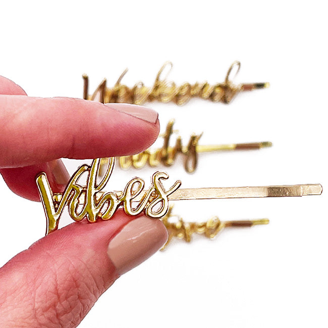 Hand with light pink nail polish holding gold On The Go barrettes, with “Vibes” in front