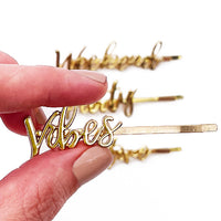 Hand with light pink nail polish holding gold On The Go barrettes, with “Vibes” in front