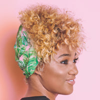 Woman with curly blonde hair shown in profile against a pink background, wearing the wide tropical leaf print headband with her curls piled on top